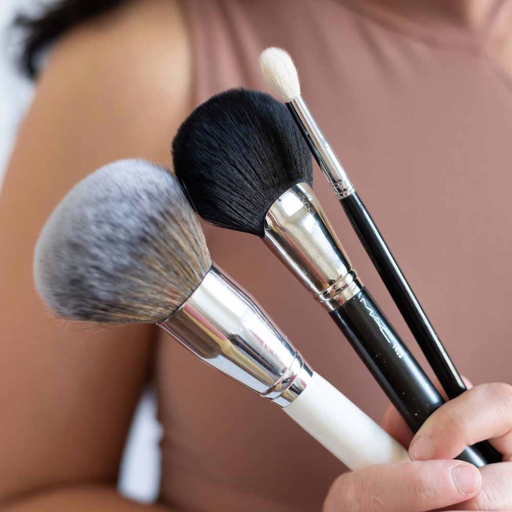 This is a branding photo taken by Vanessa Schwinn, photography in Oahu, Hawaii makeup artist Savannah Phillips. The photo is a detail shot of three make up brushes, the first one being white the second to being black in variety of sizes up against a woman's torso you can see her brown top and top of her arm.