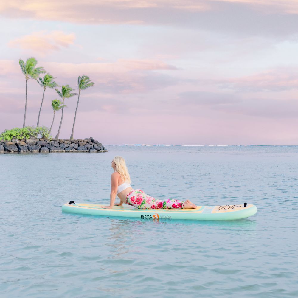 Branding photo taken by Vanessa Schwinn photography of a sup yoga, instructor student of Island paddler bliss in Honolulu Hawaii. This photo is of a woman in the ocean on a teal paddleboard doing a cobra pose. There are palm trees in the background, cotton candy clouds, and she is wearing a white top with pink floral pants and has blonde hair.
