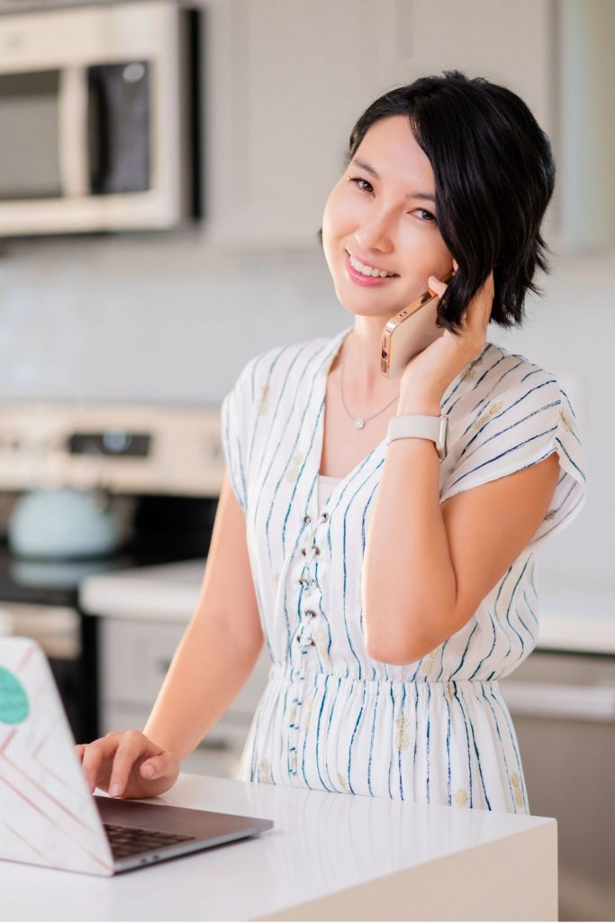 This is a branding photo taken by Vanessa Schwinn photography. The photo is of a realtor in a light and bright kitchen, standing at the counter on her laptop and talking on the phone. She is wearing a white dress and looking at the camera.