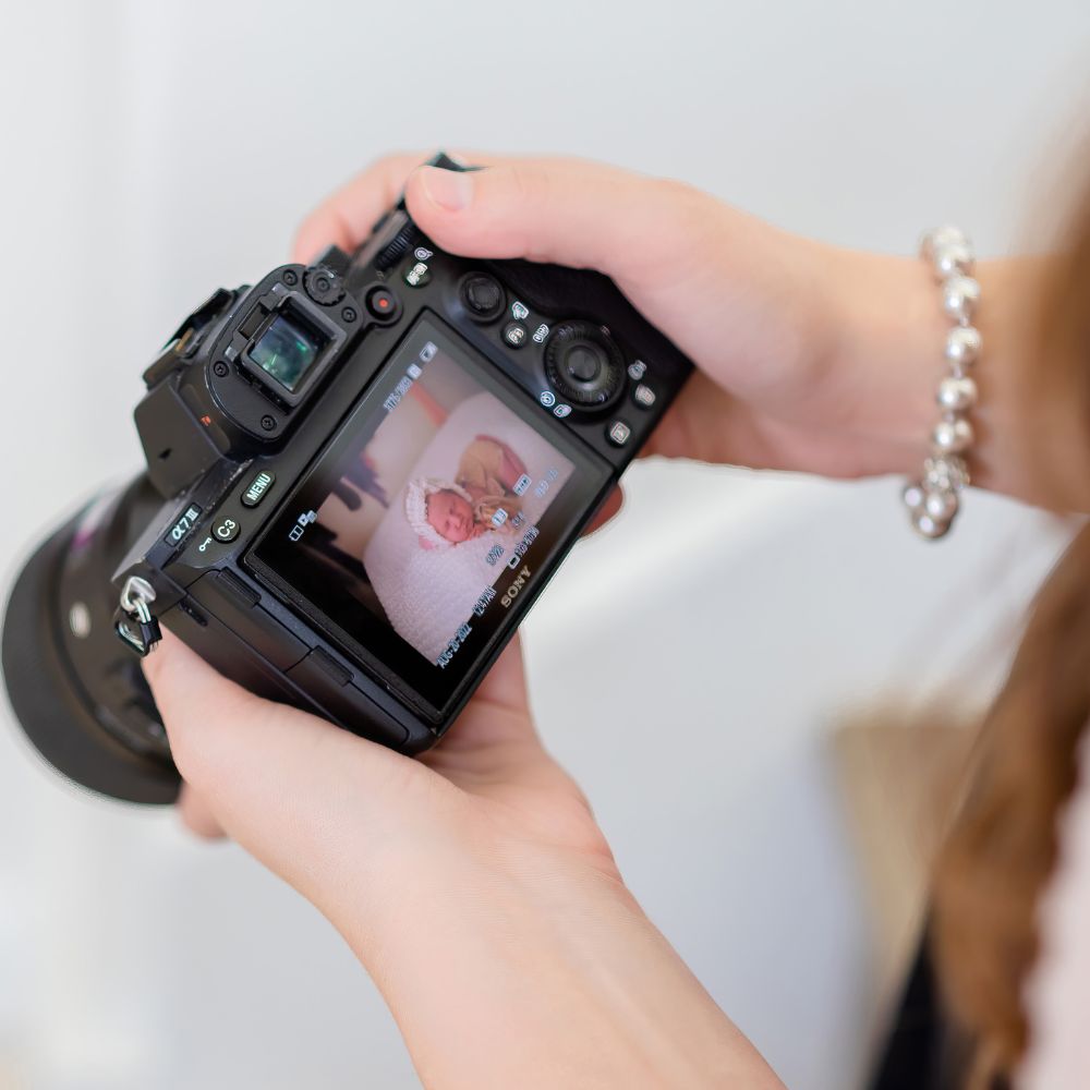 This is a photo taken by Vanessa Schwinn photography of photography by Emma Brown in Oahu, Hawaii. In the photo there is a female holding a camera with a picture of a baby on it. She has a pearl bracelet on the background is mostly white and the only thing in focus is the camera. You can see her hands and a little bit of her hair and torso.