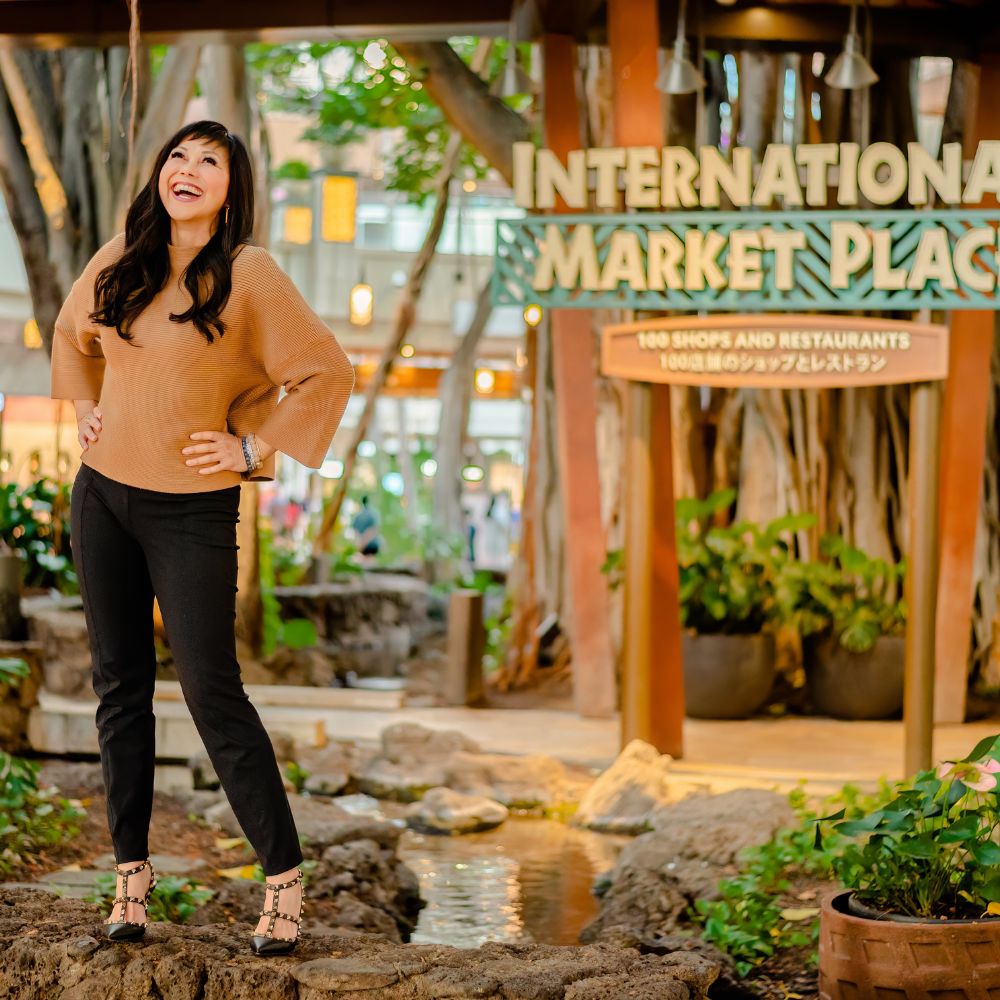 This is a branding photo shot by Vanessa Schwinn photography in Honolulu Hawaii at International Market Pl., of realtor Caron Ling. The photo is of a woman, laughing and standing with her hands on her hips and high heels on a rock in front of the International Market Place treehouse sign. The area is very wooden and has a lot of greenery.